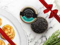 American White Sturgeon Caviar Tin Opened on Marble Surface with Creme Fraiche and Blinis with Gift Bow Graphic on Top Right