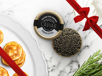 Paddlefish Caviar Tin Opened on Marble Surface with Creme Fraiche and Blinis with Gift Bow Graphic on Top Right