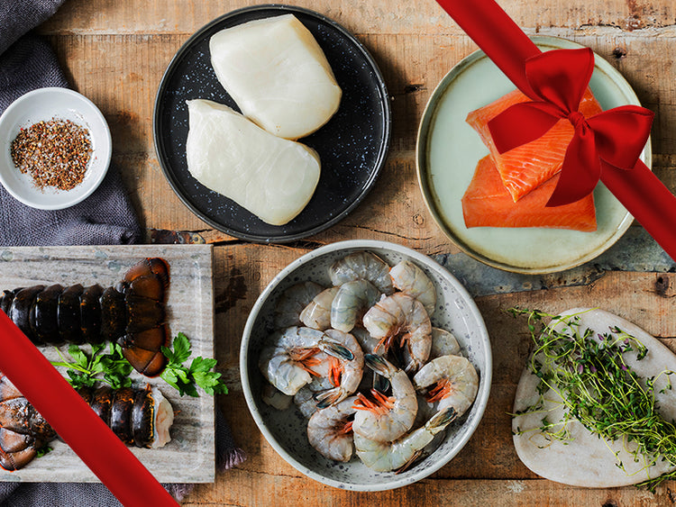 Lobster Tails, Chilean Sea Bass Portions, Wild Alaska Sockeye Salmon Portions, and Wild Blue Shrimp on Table with Seasonings and Herbs with Gift Bow Graphic on Top Right