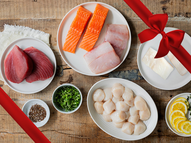 Sea Scallops, Atlantic Halibut Portions, Red Snapper Portions, Atlantic Salmon Portions, and Ahi Tuna Steaks on Plates on Wood Table with Gift Bow Graphic on Top Right