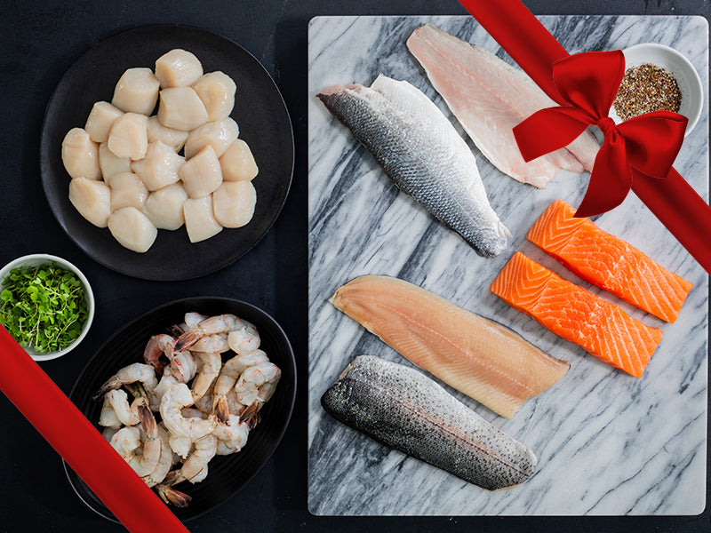 Sea Scallops and Wild Blue Shrimp on Plates and Atlantic Salmon Portions, Rainbow Trout Fillets, and Branzino Fillets on Marble Tray with Gift Bow Graphic on Top Right