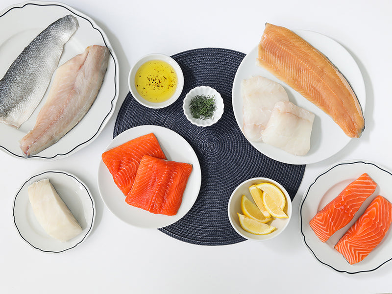 Atlantic Salmon, Arctic Char, Atlantic Cod, and Chilean Sea Bass Portions, and Branzino, and Rainbow Trout Fillets on Plates with Lemon Slices and Herbs