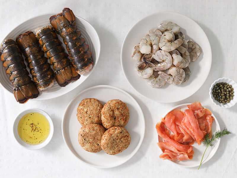 Assorted Seafood on Dishes including Lobster Tails, Shrimp, Smoked Salmon, and Crab Cakes on a White Surface