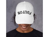 White cap with 'FULTON' text worn by a person against a gray background