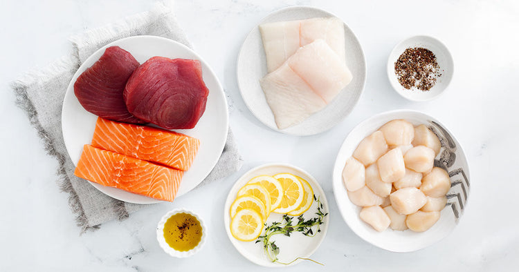 Salmon, Halibut, Tuna Steaks, and Scallops on Plates with Lemons, Herbs, and Seasonings on Marble Surface