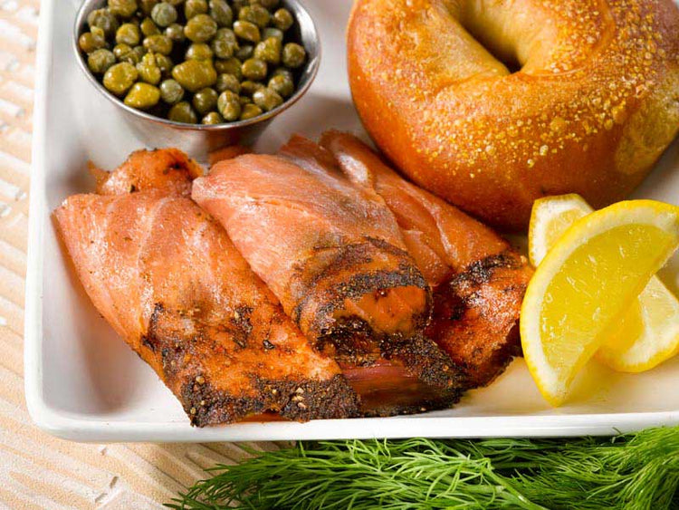 Plated pastrami smoked salmon with capers, bagel, and lemon wedges on a wooden surface