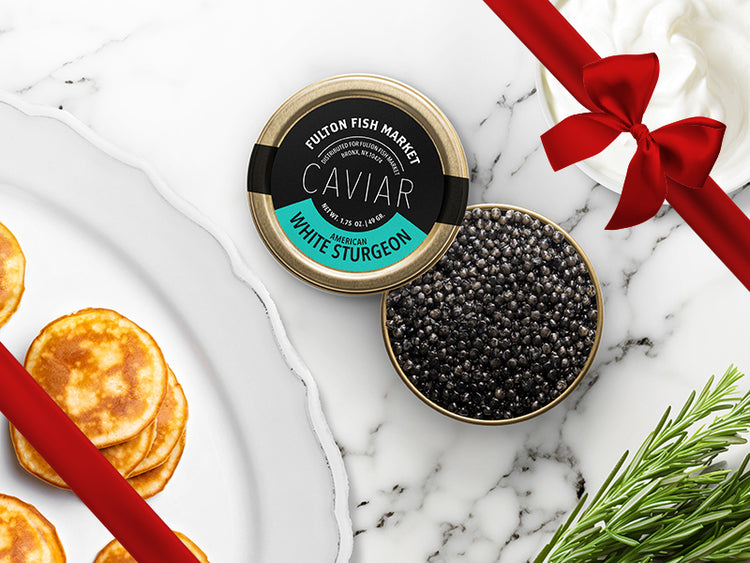 American White Sturgeon Caviar Tin Opened on Marble Surface with Creme Fraiche and Blinis with Gift Bow Graphic on Top Right