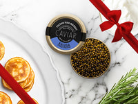 Kaluga Hybrid Strugeon Caviar Tin Opened on Marble Surface with Creme Fraiche and Blinis with Gift Bow Graphic on Top Right