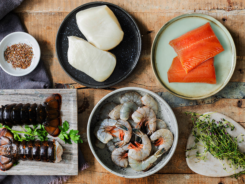 Lobster Tails, Chilean Sea Bass Portions, Wild Alaska Sockeye Salmon Portions, and Wild Blue Shrimp on Table with Seasonings and Herbs