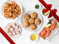 Grilled shrimp, bacon-wrapped scallops, crab cakes, smoked salmon with limes and seasonings on a marble background and a red gift bow.