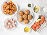 Grilled Shrimp, bacon wrapped scallops, crab cakes, smoked salmon with lemon, limes and seasonings on a white marble surface