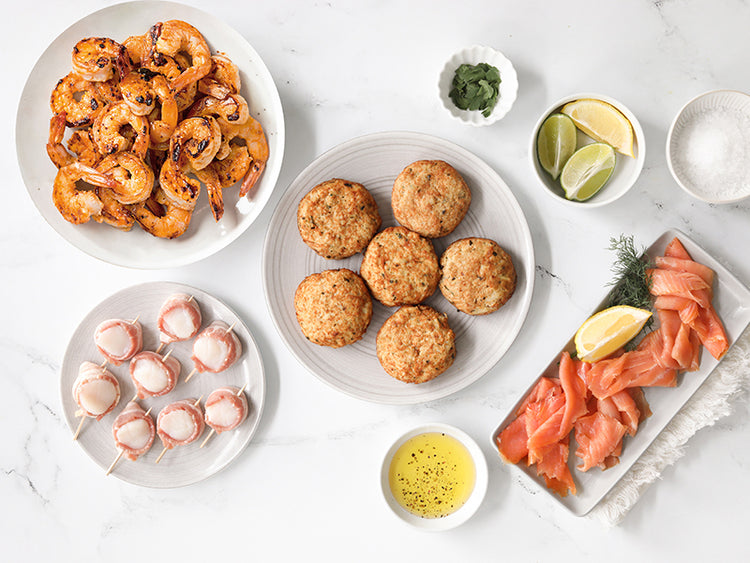 Grilled Shrimp, bacon wrapped scallops, crab cakes, smoked salmon with lemon, limes and seasonings on a white marble surface