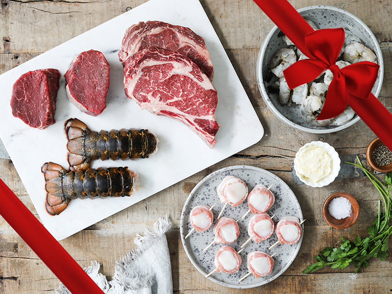 Bacon-Wrapped Scallops, Shrimp, Lobster Tails, Filet Mignon, and Ribeye Steak on Plates on Wood Table with Gift Bow Graphic