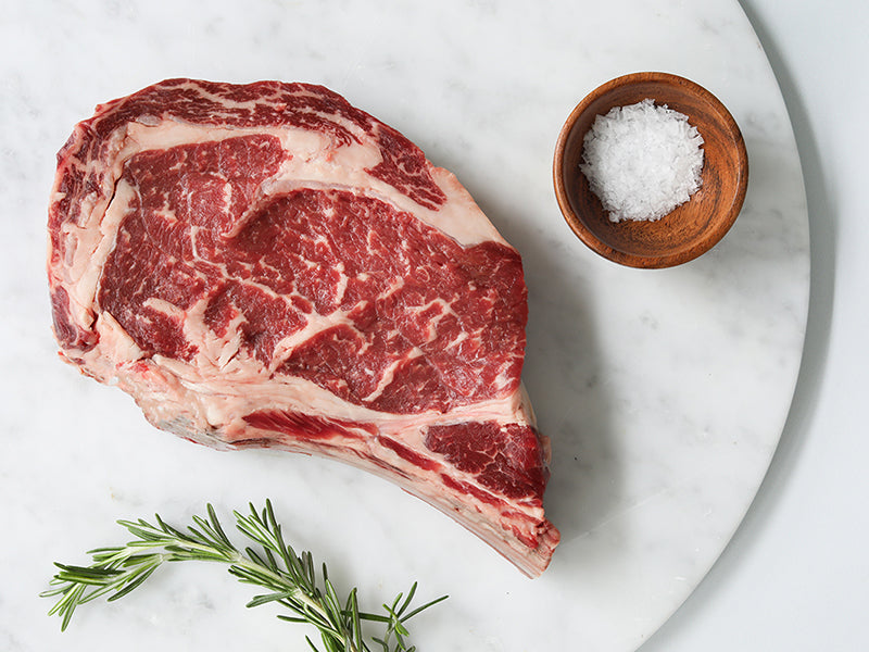 Raw Pat LaFrieda Dry Aged Ribeye Bone-In Steak with Salt and Rosemary on a Marble Surface
