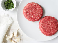 Two Raw Pat LaFrieda Dry-Aged Burger Patties on a white plate with a small bowl of parsley and garlic cloves on a white surface.