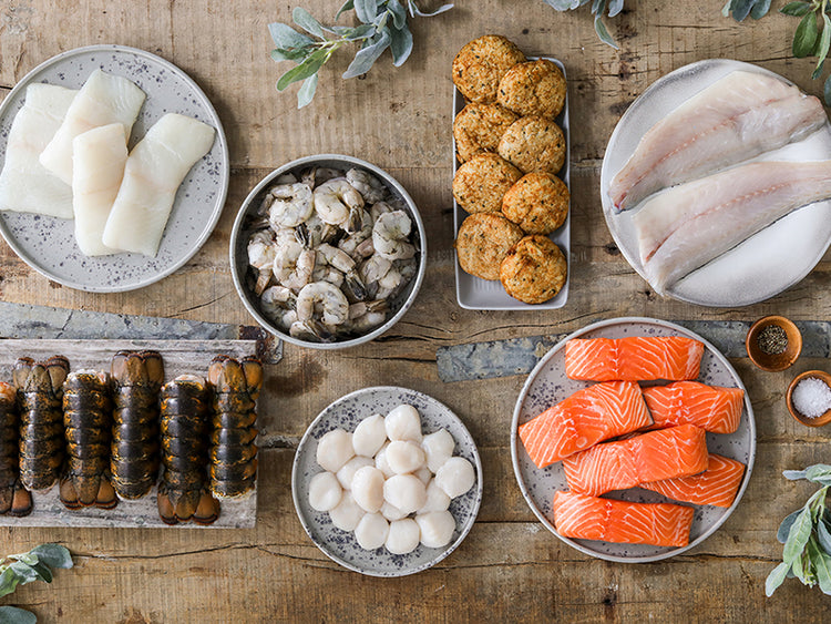 Atlantic Salmon Portions, Wild Cold Water Lobster Tails, Scallops, Crab Cakes, Atlantic Halibut Portions, and Branzino Fillets on Plates on Wooden Table with Holiday Greenery