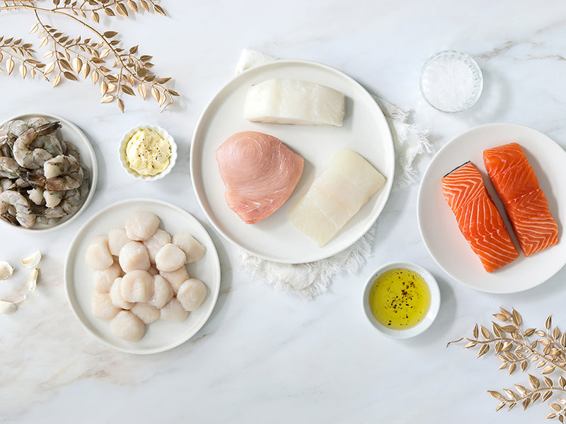 Holiday Sampler Bundle of Scallops, Shrimp, Atlantic Salmon Portions, Chilean Sea Bass Portion, Swordfish Steak, and Atlantic Halibut Portion on Plates with Festive Gold Leaves