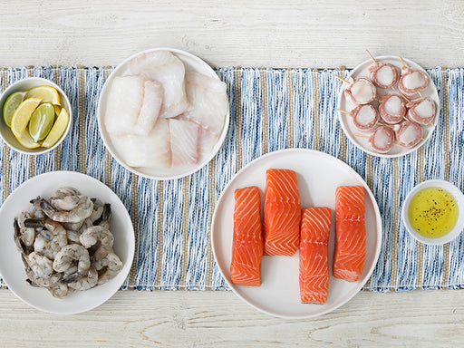 Shrimp, Atlantic Salmon and Atlantic Cod Portions, and Bacon-Wrapped Scallops on Striped Tablecloth with Lemon and Lime Wedges