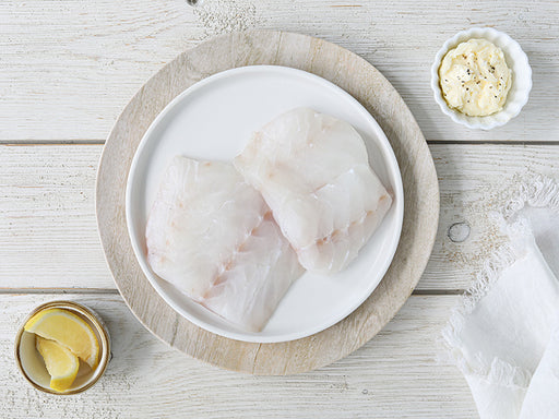 Two Portions of Atlantic Cod on Plate on Light Wood Table with Lemon Wedges and Compound Butter