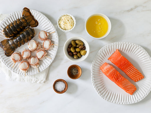 Lobster Tails, King Salmon Portions, and Bacon-Wrapped Scallops on Plates with Compound Butter, Olive Oil, Seasonings, and Capers