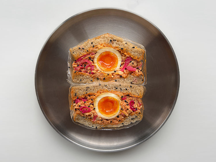 Two halves of a sandwich  on a round metal plate, filled with Fishwife Albacore Tuna in Olive Oil and a halved soft-boiled egg on a light gray background