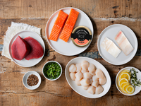 Ahi Tuna, Atlantic Salmon, Halibut, Salmon Roe tin, and scallops on white plates with herbs, spices and sliced lemons on a wood table. 