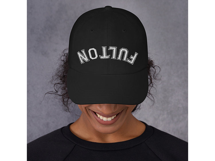 Person wearing a black cap with 'FULTON' embroidered on it against a gray background