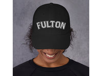 Person wearing a black cap with 'FULTON' embroidered on it against a gray background