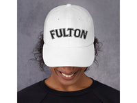 White cap with 'FULTON' text worn by a person against a gray background