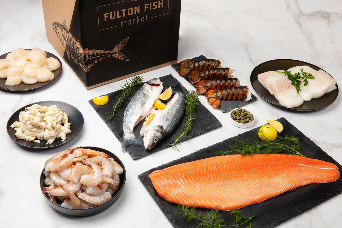 Fulton Fish Market Box on Marble Surface with Shrimp, Crabmeat, Scallops, Butterflied Branzino, Lobster Tails, and Salmon Fillet on Plates with Lemons and Herbs